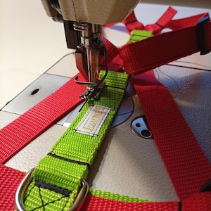 Könnte beinhalten: Nahaufnahme einer Nähmaschine, die ein rotes und grünes Hundegeschirr näht. Das Geschirr besteht aus strapazierfähigem Nylongewebe mit einem Metall-D-Ring und einem Etikett mit der Aufschrift "Journey". Die Nadel der Maschine befestigt einen grünen Gurt an einem roten Gurt.