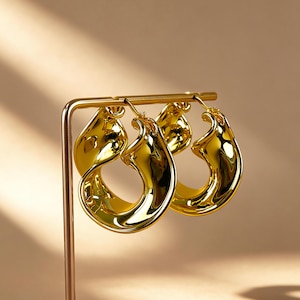 May include: A pair of gold-colored hoop earrings with a unique, organic shape. The earrings are displayed on a gold-colored stand against a neutral background. The earrings have a glossy finish, reflecting light.