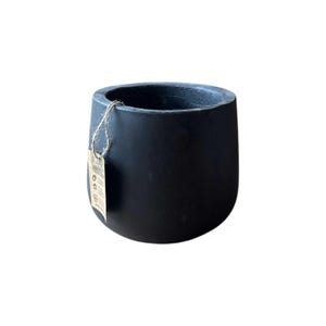 Puede incluir: Una maceta redondeada de color negro mate con una parte superior ancha y abierta. Una pequeña etiqueta de papel beige rectangular está unida al borde de la maceta con un trozo fino de cuerda de yute natural. La superficie es lisa y uniforme.