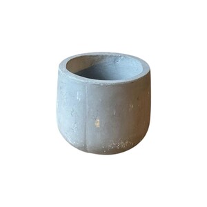 Puede incluir: Una maceta cilíndrica de color gris con un acabado mate suave y una base ligeramente redondeada. La superficie exterior presenta sutiles variaciones de tono y textura, con una costura vertical visible a lo largo del lado del recipiente.