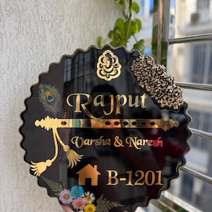 May include: Black, scalloped-edge nameplate with gold lettering and accents. The name "Rajput" is displayed prominently, with "Varsha & Naresh" and the address "B-1201" below. Features a peacock feather, floral and gold embellishments.