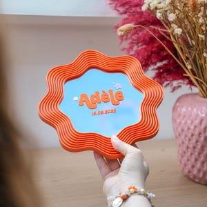 May include: A wavy, orange, flower-shaped mirror featuring the name Adèle in bold orange letters, accompanied by two small white flower icons and the date 18.09.2025 printed in orange on the reflective surface of the mirror.