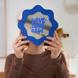 May include: A blue wavy-edged mirror with the words "LOVE YOUR SELF" in blue and white. The mirror is held up in front of a person. The mirror is a decorative item.