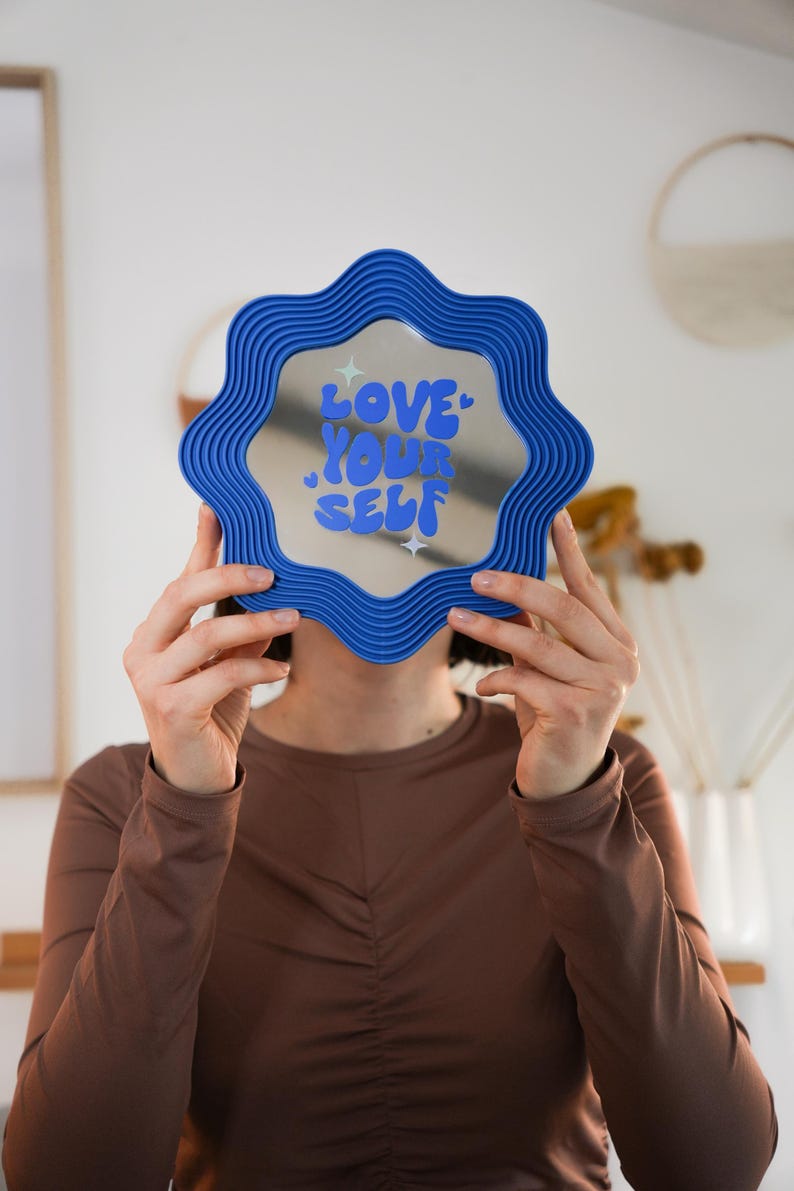 May include: A blue wavy-edged mirror with the words "LOVE YOUR SELF" in blue and white. The mirror is held up in front of a person. The mirror is a decorative item.