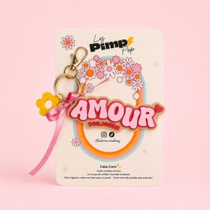 May include: A keychain with a pink and red "AMOUR TOUJOURS" design, a yellow flower charm, and a pink ribbon. The keychain is attached to a card with the text "Les Pimp Pop" and a floral design.