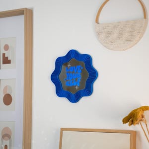 May include: A blue wavy-edged mirror with the text "LOVE YOUR SELF" in blue lettering. The mirror is mounted on a white wall, alongside framed artwork and a woven wall hanging. Decorative home decor.