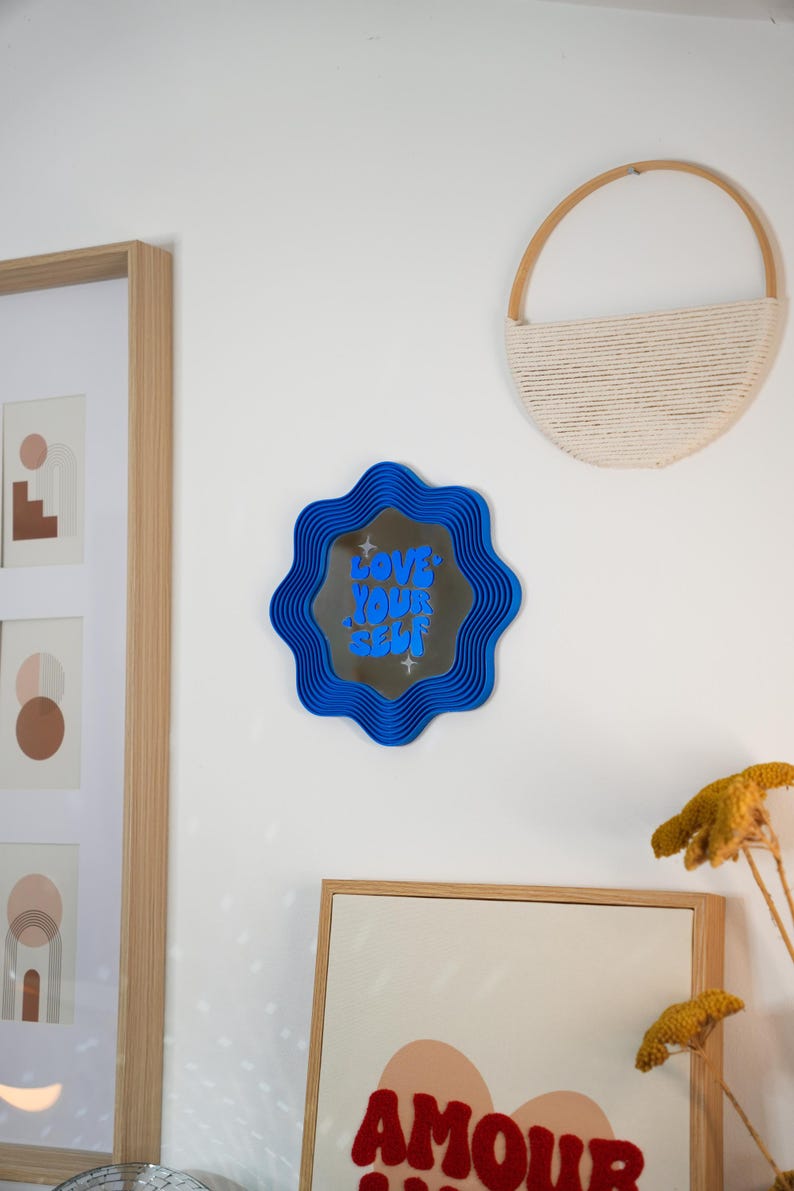 May include: A blue wavy-edged mirror with the text "LOVE YOUR SELF" in blue lettering. The mirror is mounted on a white wall, alongside framed artwork and a woven wall hanging. Decorative home decor.