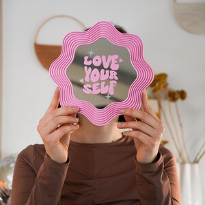 May include: A pink, wavy-edged mirror with the words "LOVE YOUR SELF" in white script. The mirror is held up in front of a person's face. The background is a light pink wall with decorative items.
