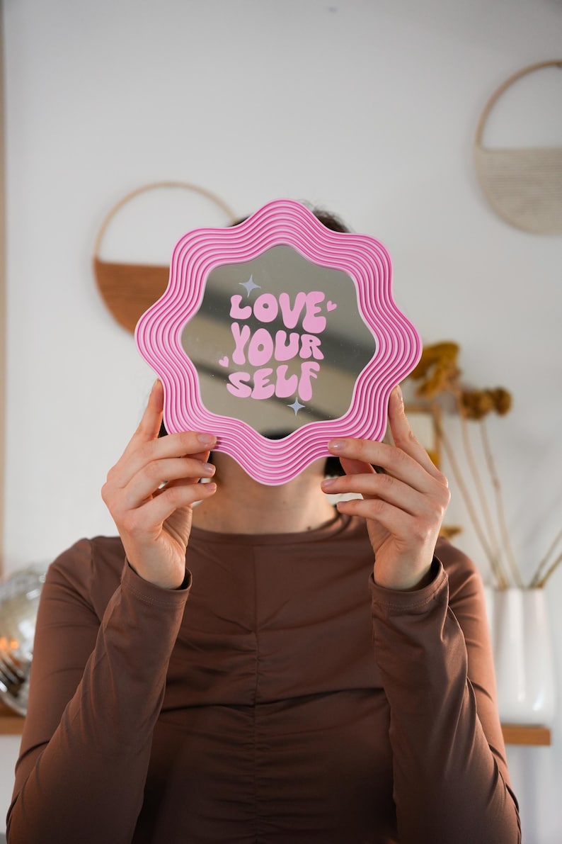 May include: A pink, wavy-edged mirror with the words "LOVE YOUR SELF" in white script. The mirror is held up in front of a person's face. The background is a light pink wall with decorative items.