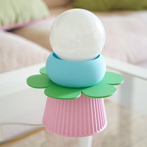 Peut inclure: Une lampe de table composée d'une ampoule sphérique blanche reposant sur un anneau bleu clair texturé, une base verte en forme de fleur et un support rose cannelé style cupcake avec un cordon d'alimentation blanc sortant du côté.