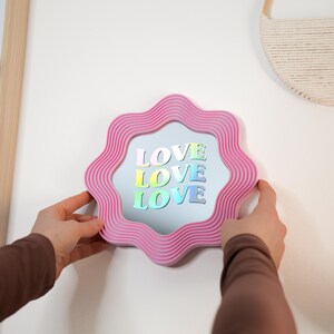 May include: A pink, wavy-edged mirror with the word "LOVE" in colorful, holographic letters. The mirror is being held up against a white wall. A decorative wall hanging and a framed picture are also visible.