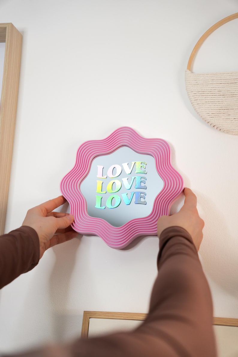 May include: A pink, wavy-edged mirror with the word "LOVE" in colorful, holographic letters. The mirror is being held up against a white wall. A decorative wall hanging and a framed picture are also visible.