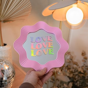 以下が含まれることがあります： ホログラフィック文字で「LOVE」と書かれた、ピンク色の波型の縁取りの鏡。 鏡はニュートラルな背景の前に保持され、ディスコボールと花型のランプが見えます。 鏡は装飾品です。