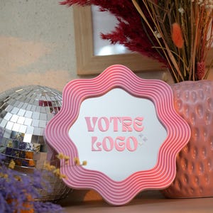 May include: A pink, wavy-edged mirror with the text "VOTRE LOGO" in pink. The mirror is displayed with a disco ball, dried flowers, and a pink vase. The overall theme is decorative and stylish.