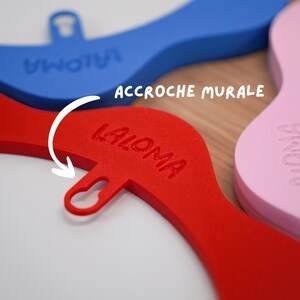 May include: Close-up of colorful wall hooks in red, blue, and pink. The red hook has the word "LALOMA" embossed on it, and the text "ACCROCHE MURALE" is visible. The hooks are designed with a curved shape and a loop for hanging.