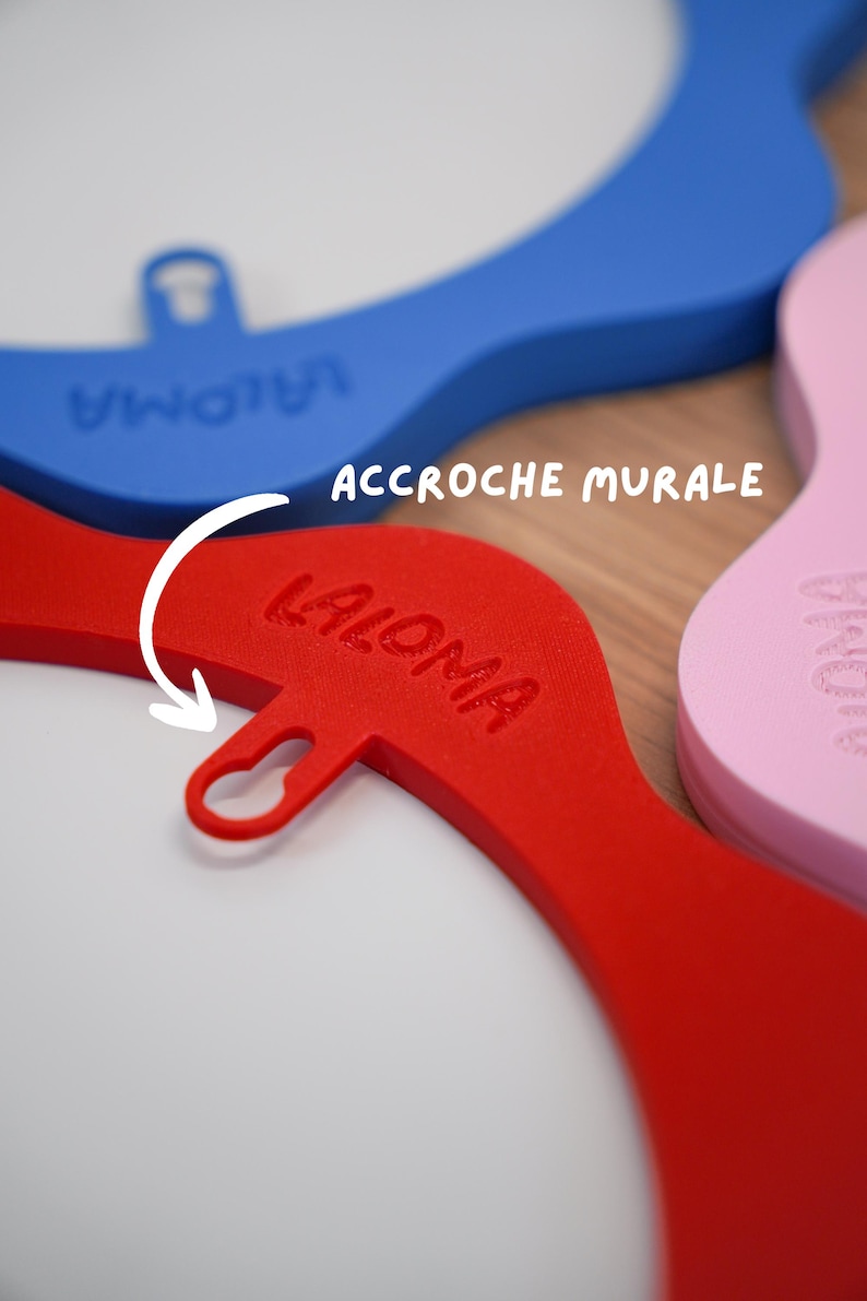 May include: Close-up of colorful wall hooks in red, blue, and pink. The red hook has the word "LALOMA" embossed on it, and the text "ACCROCHE MURALE" is visible. The hooks are designed with a curved shape and a loop for hanging.