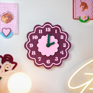 Peut inclure: Une horloge murale festonnée avec une base violet foncé, un centre rose clair et des chiffres rose clair assortis de un à douze. L'horloge possède deux aiguilles vert foncé et un design de bordure ondulé en forme de fleur.