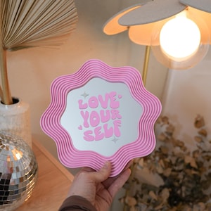 May include: A pink wavy-edged mirror with the words "LOVE YOUR SELF" in pink lettering. The mirror is held in front of a neutral background with a lamp and decorative items.