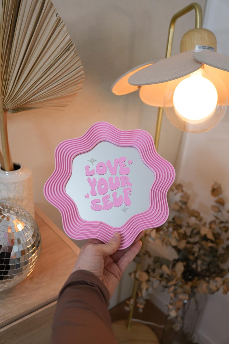 May include: A pink wavy-edged mirror with the words "LOVE YOUR SELF" in pink lettering. The mirror is held in front of a neutral background with a lamp and decorative items.