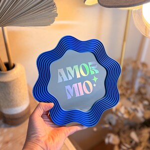 May include: A blue, wavy-edged mirror with the words "AMOR MIO" in iridescent lettering. The mirror is held up in front of a neutral background. The mirror has a unique, decorative design.