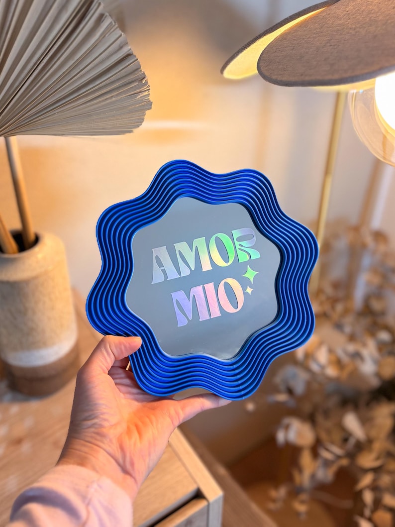 May include: A blue, wavy-edged mirror with the words "AMOR MIO" in iridescent lettering. The mirror is held up in front of a neutral background. The mirror has a unique, decorative design.