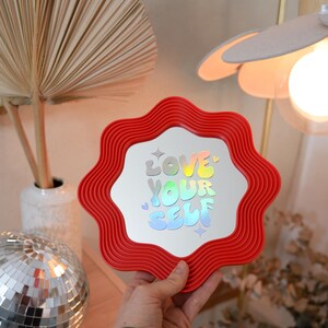May include: A red, wavy-edged mirror with the words "LOVE YOUR SELF" in holographic lettering. The mirror is held in front of a neutral background with a disco ball and a decorative lamp.
