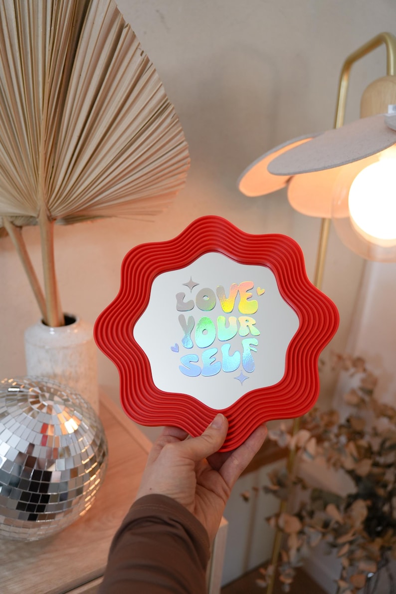 May include: A red, wavy-edged mirror with the words "LOVE YOUR SELF" in holographic lettering. The mirror is held in front of a neutral background with a disco ball and a decorative lamp.