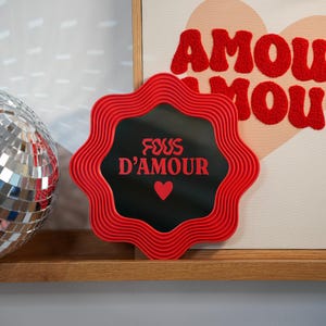 May include: A red, wavy-edged mirror with the text "FOUS D'AMOUR" and a heart symbol. The mirror is set against a black background. A disco ball and a framed artwork with the word "AMOUR" are also visible.