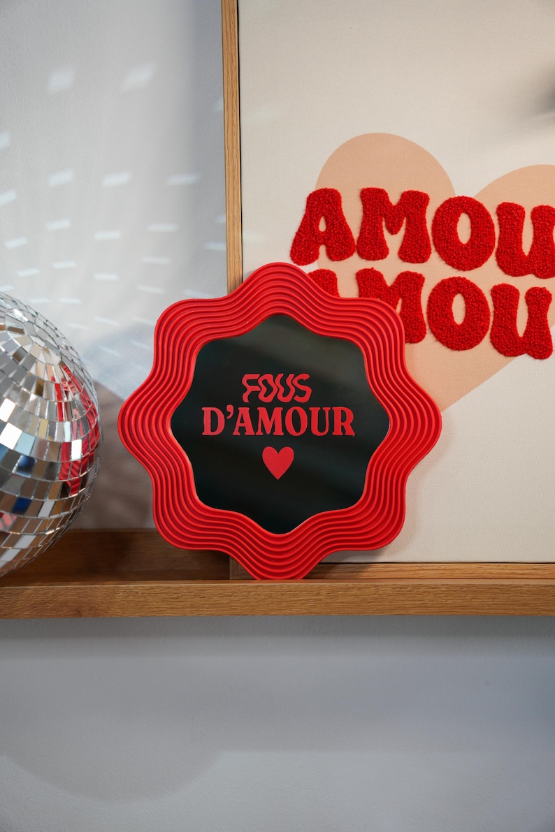 May include: A red, wavy-edged mirror with the text "FOUS D'AMOUR" and a heart symbol. The mirror is set against a black background. A disco ball and a framed artwork with the word "AMOUR" are also visible.