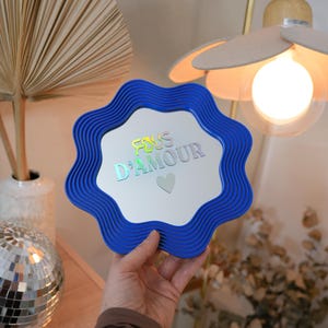 May include: A blue, wavy-edged mirror with the text "FOUS D'AMOUR" in iridescent lettering and a small heart. The mirror is held up in front of a light fixture and a decorative arrangement.