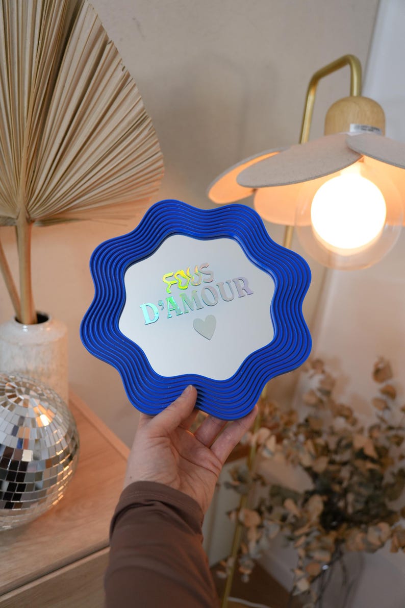 May include: A blue, wavy-edged mirror with the text "FOUS D'AMOUR" in iridescent lettering and a small heart. The mirror is held up in front of a light fixture and a decorative arrangement.
