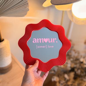 May include: A red, wavy-edged mirror with the word "amour" in pink, and the word "love" in brackets. The mirror is held up in front of a light-colored background. The mirror's frame has a textured, ribbed design.