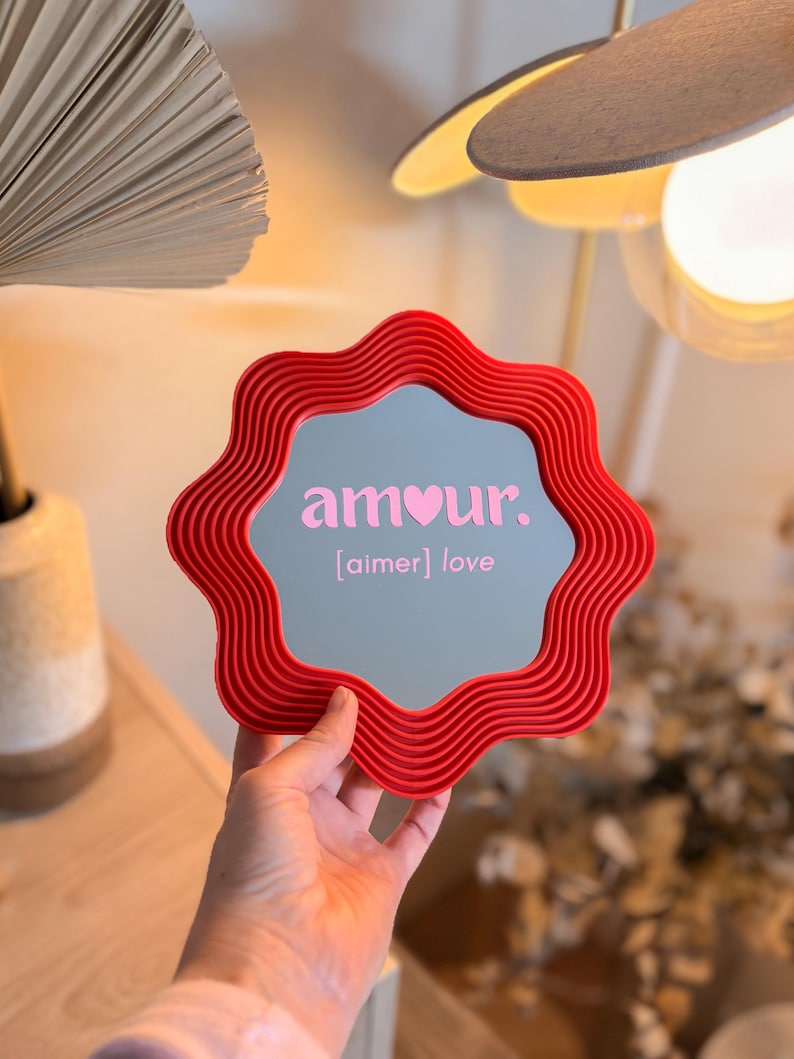 May include: A red, wavy-edged mirror with the word "amour" in pink, and the word "love" in brackets. The mirror is held up in front of a light-colored background. The mirror's frame has a textured, ribbed design.