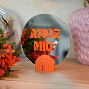 Peut inclure: Un miroir rond avec les mots "AMOR MIO" en orange, posé sur une surface en bois. Un petit objet orange en forme d'arc-en-ciel est devant le miroir. Le miroir reflète des fleurs oranges et une boule à facettes.