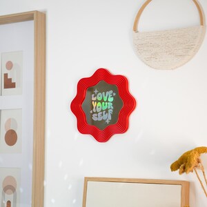 May include: A red, wavy-edged mirror with the words "LOVE YOUR SELF" in holographic lettering. The mirror is mounted on a white wall, alongside framed art and a woven wall hanging. The overall aesthetic is modern and decorative.