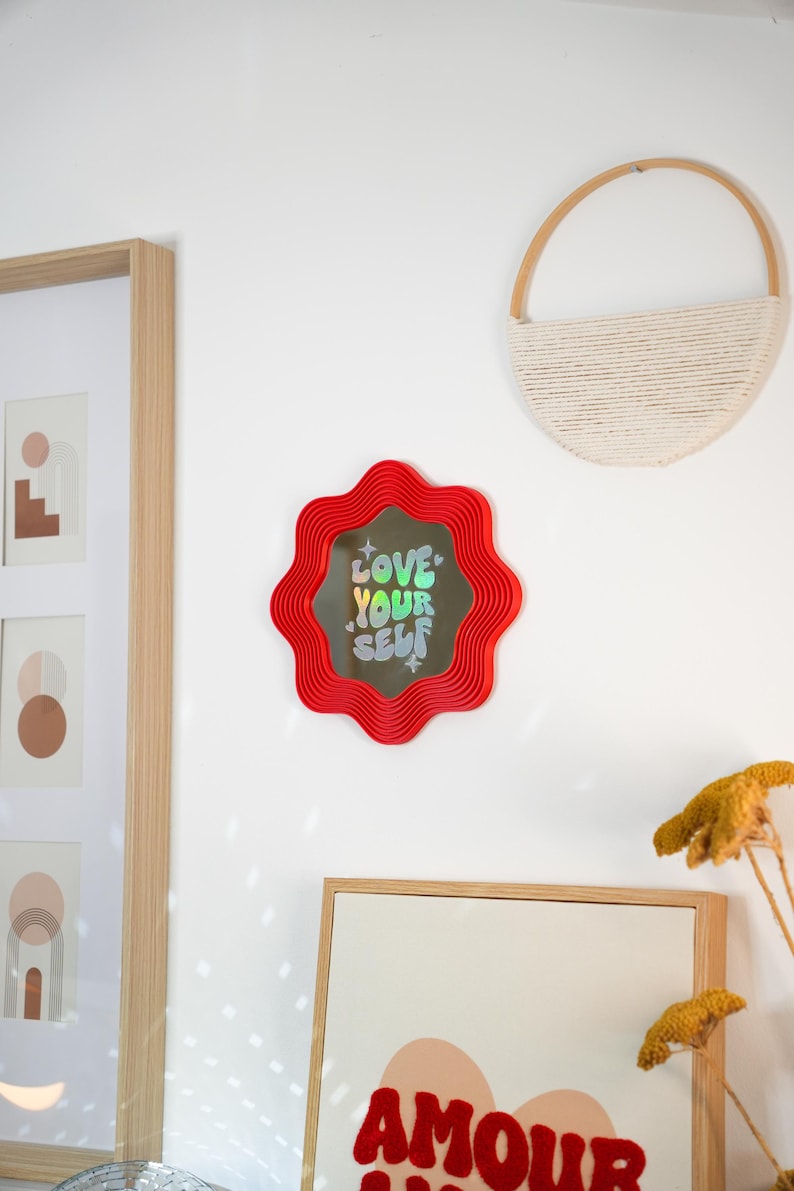 May include: A red, wavy-edged mirror with the words "LOVE YOUR SELF" in holographic lettering. The mirror is mounted on a white wall, alongside framed art and a woven wall hanging. The overall aesthetic is modern and decorative.