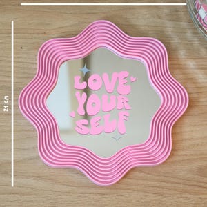 May include: A pink, wavy-edged mirror with the text "LOVE YOUR SELF" in bubble letters. The mirror is 21 cm (8.3 inches) wide and 21 cm (8.3 inches) tall. The frame has a ribbed design.