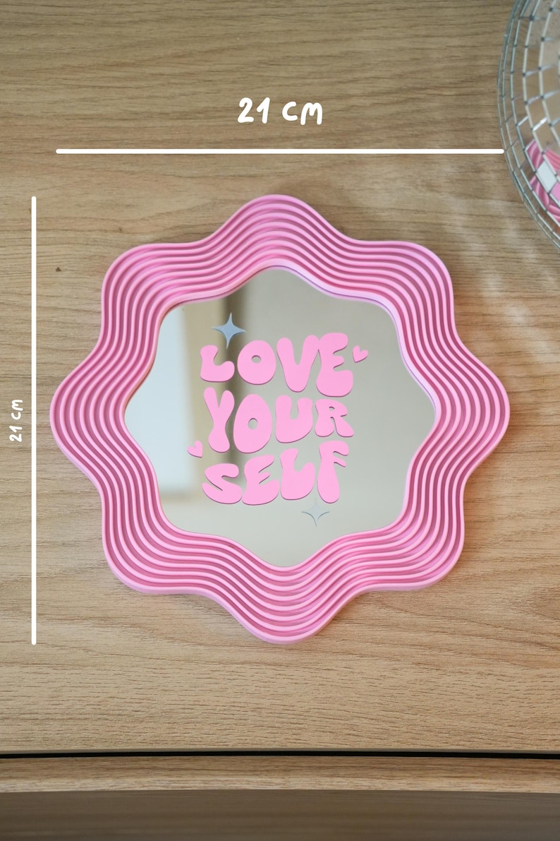 May include: A pink, wavy-edged mirror with the text "LOVE YOUR SELF" in bubble letters. The mirror is 21 cm (8.3 inches) wide and 21 cm (8.3 inches) tall. The frame has a ribbed design.