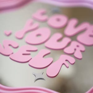 May include: A pink decorative tray with the words "LOVE YOUR SELF" in bubble letters. The tray has a wavy, ridged border. A small star graphic is below the text. The background is a light neutral color.