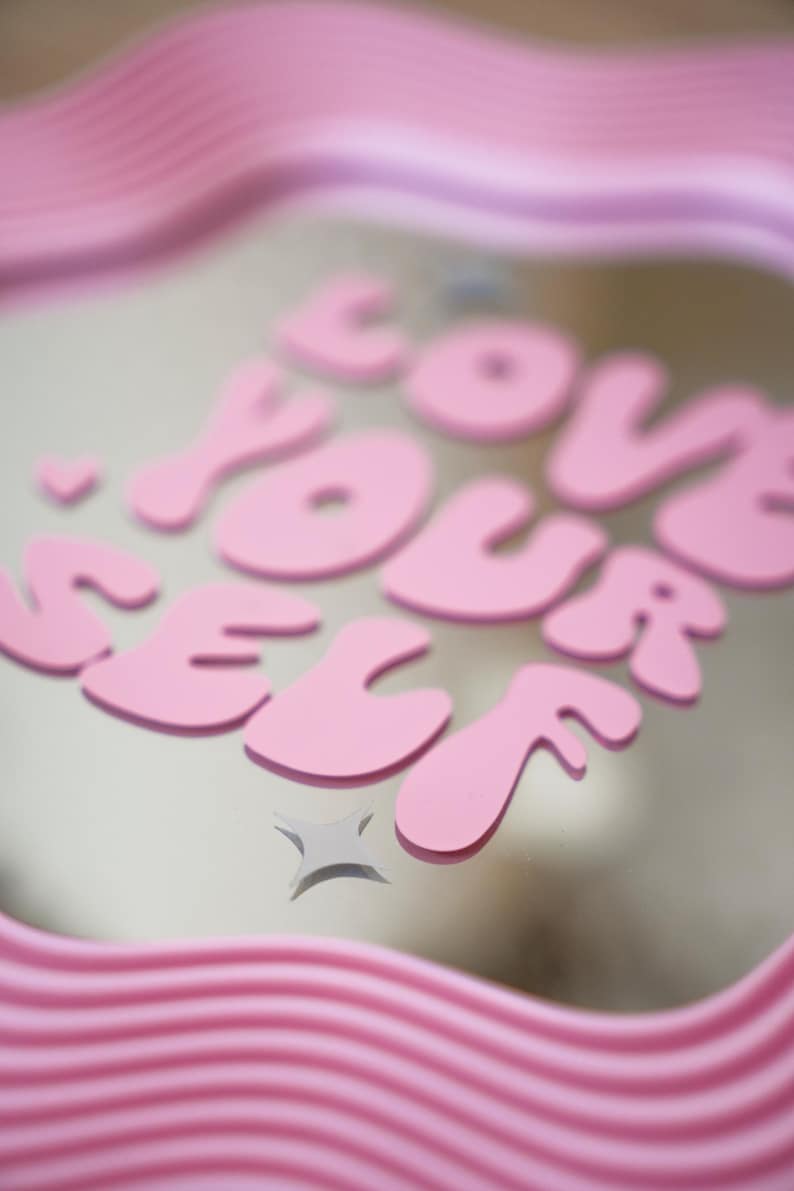 May include: A pink decorative tray with the words "LOVE YOUR SELF" in bubble letters. The tray has a wavy, ridged border. A small star graphic is below the text. The background is a light neutral color.