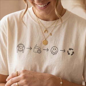 Puede incluir: Camiseta color crema con un gráfico negro que representa una secuencia de imágenes: un volante, un altavoz, una carita sonriente y un símbolo de reciclaje. También se ven collares y anillos dorados.