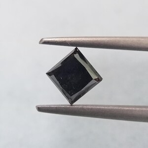 Op de afbeelding: Een zwarte diamant in princess-slijpvorm met een vierkante vorm en een gefacetteerd oppervlak. De edelsteen wordt vastgehouden door een metalen pincet en toont zijn donkere, ondoorzichtige kleur en scherpe geometrische randen tegen een lichte achtergrond.