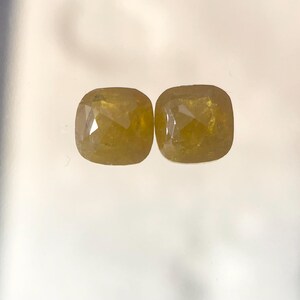 May include: Two cushion-cut yellow gemstones with faceted surfaces. These loose stones exhibit a deep golden hue and a symmetrical, rounded square shape, reflecting light through their polished, multi-faceted tops against a plain background.
