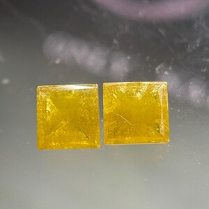 May include: Two square-cut yellow gemstones with faceted surfaces. Each stone displays a vibrant, translucent golden-yellow hue with internal inclusions and a sharp, geometric princess-cut shape that reflects light across its flat top surface.