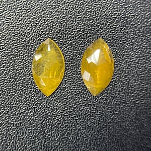 May include: Two marquise-cut yellow gemstones with faceted surfaces. Each stone features a pointed oval shape with a vibrant golden hue and multiple reflective facets that catch the light, displaying a translucent quality and a polished finish.