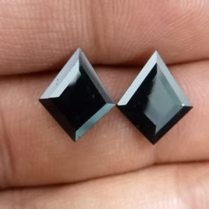 May include: Two loose, kite-shaped black gemstones with a faceted surface. Each stone features a pointed top and bottom with angled sides, creating a geometric silhouette. The stones have a smooth, polished finish that reflects light across their dark surfaces.