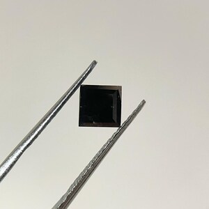 Op de afbeelding: Een vierkant geslepen, donkerzwarte edelsteen vastgehouden door een zilverkleurige metalen pincet. De steen heeft scherpe, strakke randen en een plat, gefacetteerd oppervlak dat licht reflecteert en een diepe, ondoorzichtige kleur laat zien.