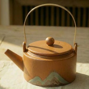 Könnte beinhalten: Eine braune Keramik-Teekanne mit Deckel und gebogenem Griff. Die Teekanne hat einen Ausguss und einen kleinen Knopf auf dem Deckel. Der Boden der Teekanne hat ein dekoratives grün-weißes Bergdesign.