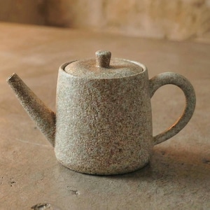 Op de afbeelding: Een kleine, bespikkelde keramische theepot met een deksel en een gebogen handvat. De theepot heeft een tuit en een neutraal kleurenpalet, geschikt voor het serveren van thee. Het ontwerp is eenvoudig en rustiek, perfect voor een gezellige keuken of eetkamer.