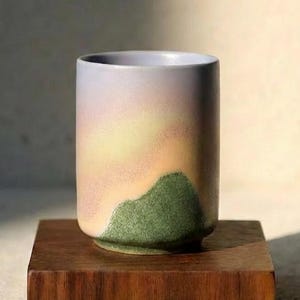 Puede incluir: Una taza cilíndrica de cerámica con un degradado de colores, incluyendo lavanda, melocotón y verde. La taza está colocada sobre un cubo de madera oscura, con un fondo neutro. El diseño de la taza se asemeja a un paisaje.
